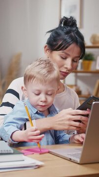 Busy Asian mother holding phone and texting while sitting with lively toddler. Child drawing energetically on pink paper. Handling online communication and parenting in same moment.