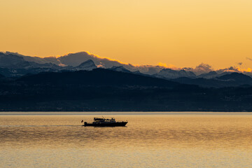 Obraz premium Boat Silhouette at Sunset with Golden Water and Mountains