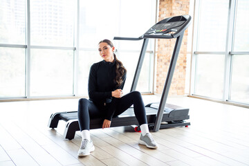 Young sportswoman resting by a treadmill in a modern gym with city views, showcasing an active and...