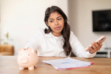 Young woman examines financial documents while using smartphone near piggy bank at home during...