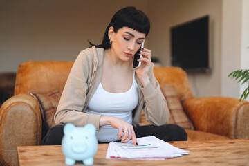 Woman on phone discussing finances while reviewing documents at home in cozy living room with...