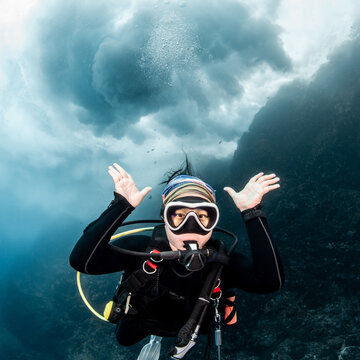 Asian female scuba diver looking at camera posing with the stunning wave breaking exploding above her head at Bon Island, Similan National Park, Thailand. Recreational underwater activity