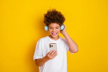 Happy boy with curly hair wearing headphones and smiling while looking at a smartphone against a...