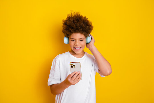 Happy boy with curly hair wearing headphones and smiling while looking at a smartphone against a yellow background