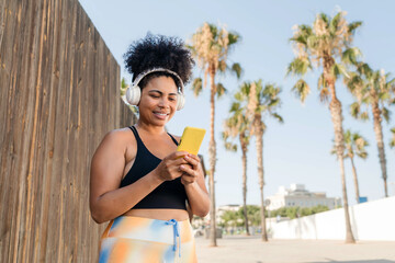 Curvy smiling Hispanic woman listening music using smartphone before workout outdoors