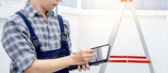 Building inspector man in workwear using digital tablet checking for the process of house project. Asian male worker writing on smart device standing with a ladder at construction site.