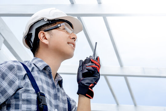 Confident Asian man engineer talking on walkie talkie while looking at roof structure. Smart foreman using radio communication equipment at construction site for cooperation with the worker team.