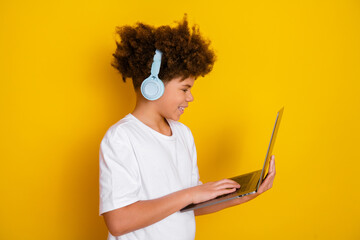 Smiling boy in white t-shirt using laptop wearing headphones against a bright yellow background...