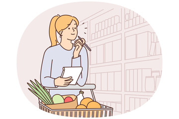 Woman walks around supermarket with cart filled with vegetables and holds checklist in hands
