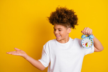 Cheerful boy holding colorful alarm clock and gesturing against yellow background showcasing time...