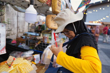 An elderly woman holds a drink and buys traditional snacks at an outdoor market stall in Pasar...