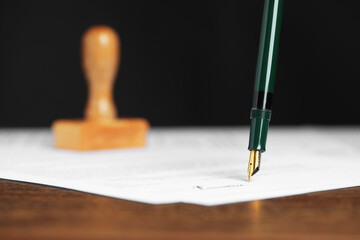 Signing notary document with fountain pen at wooden table against black background, closeup
