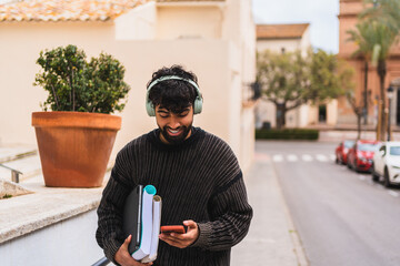 Young man walking in the city using his phone while carrying books and wearing headphones