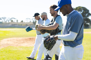 Diverse male teammates walking across sunlit grass field wearing chest protector, caps and gloves