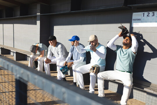 Diverse male baseball teammates sitting on bench in dugout holding bats and gloves