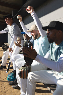 Diverse male baseball teammates cheering while sitting on bench in dugout with bat and gloves