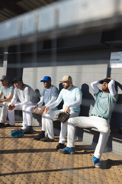 Diverse male baseball teammates sitting on bench in dugout adjusting gloves near VISITORS sign