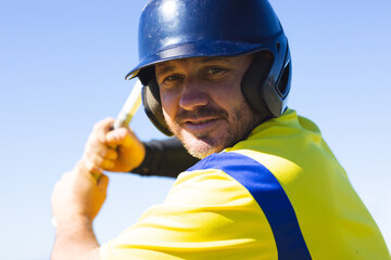 Male baseball batter wearing blue helmet and yellow jersey, swinging wooden bat on baseball field