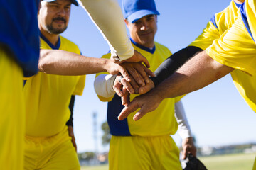 Diverse male teammates huddling on grass field with yellow jerseys caps stacking hands over glove