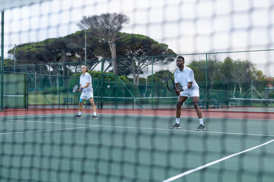 Diverse male tennis partners positioning near baseline on hard court behind net holding rackets
