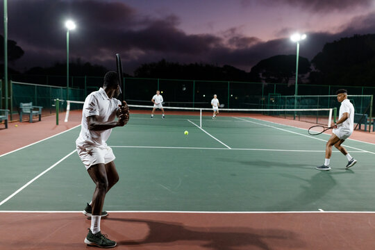 Four male players swinging rackets at neon ball under lights on court at dusk, copy space