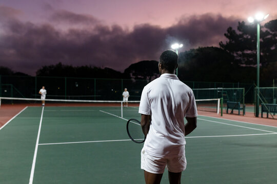 Diverse male tennis players competing on court at dusk under floodlights with rackets, net