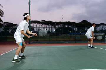 Diverse male doubles partners standing ready on tennis court at dusk with rackets, blue knee tape
