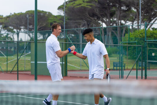 Diverse male tennis teammates exchanging celebratory fist bump at net on green court with racket