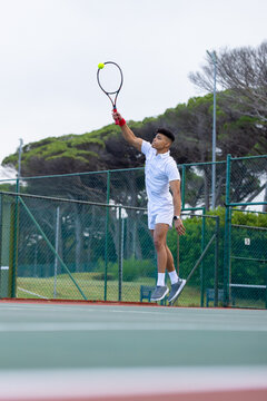 African American male tennis player jumping to serve ball on outdoor court in white holding racket