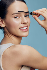 Young woman applying mascara, showcasing a natural beauty routine with a bright blue background,...