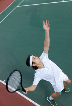 Asian male tennis player serving ball on outdoor hard court with tennis racket and white headband