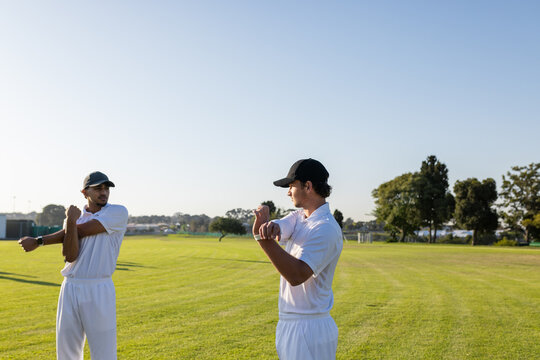 Diverse male teammates stretching across chest on cricket field in white uniforms and black caps - Powered by Adobe