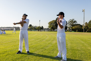 Diverse male teammates stretching arms across chest wearing white uniforms and caps on grassy field