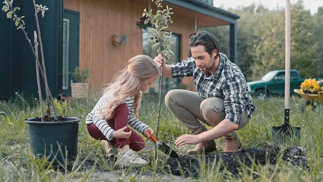 Handsome father communicating with his lovely doughter in garden. Little girl with small gardening shovel digging hole for plants while her dad telling inetisting story. Parent supporting his child.