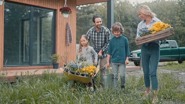 Family with gardening tools walking on green herbs in garden. People going to plant flowers. Father with his daughter riding wheelbarrow full of greenery while his wife and son walking alongside him. - Powered by Adobe