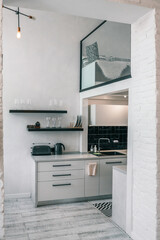 Modern minimalist kitchen with black backsplash and light wood flooring. Glass shelves hold dishes, and a cozy loft bed with pillows is visible above
