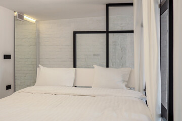 Minimalist bedroom with white bedding and industrial-style glass partition. Soft lighting and white...