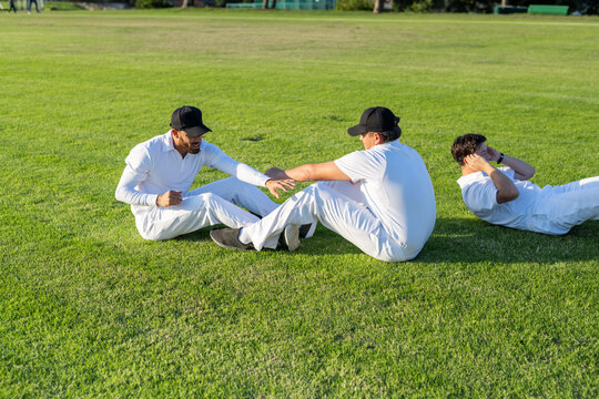 Diverse male teammates performing warm-up on cricket outfield wearing white uniforms and black caps