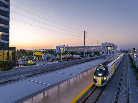 Aerial view of a sleek train glides into the station as the sun sets, casting soft hues over the modern architecture of the building, Aventura, Florida, United States.