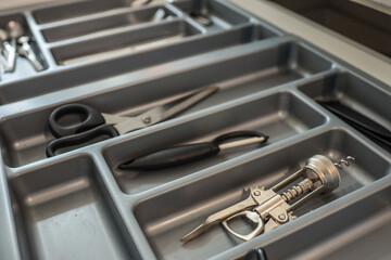 Close-up of a kitchen drawer with compartments holding scissors, a vegetable peeler, and a metal...