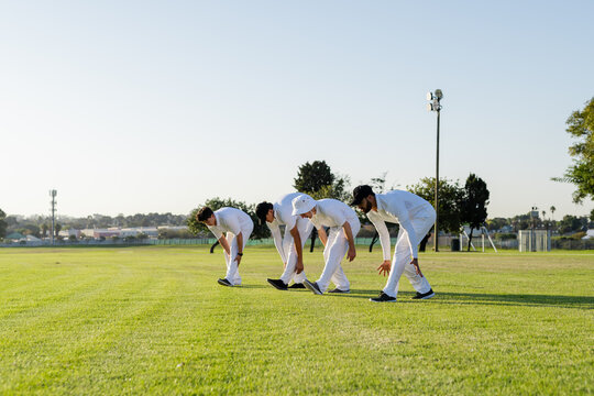 Diverse male cricket players bending forward and stretching on outfield wearing uniforms and caps - Powered by Adobe