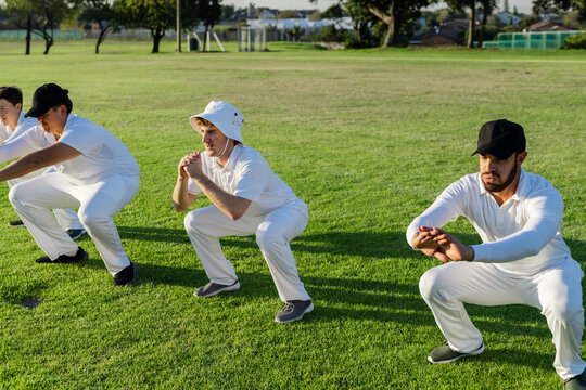 Diverse male teammates practicing catching drills on cricket field wearing white uniforms and caps