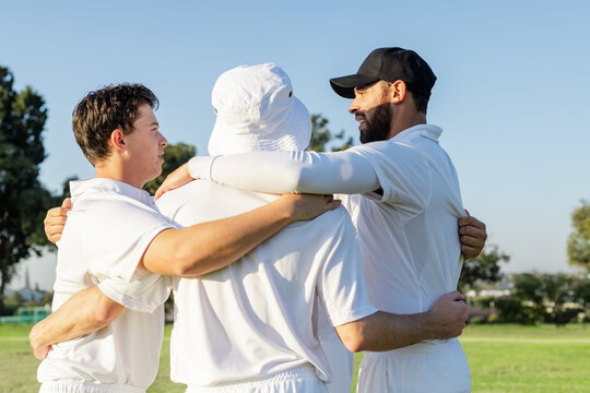 Three male teammates huddling on cricket field wearing white sports shirts, trousers and caps
