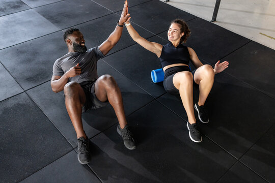 Diverse friends lying on gym floor using blue foam roller and high-fiving during crunch exercise