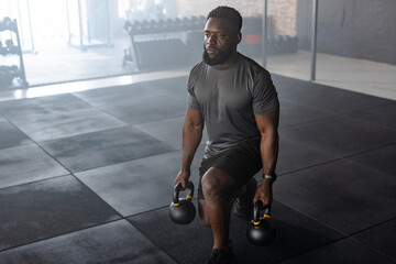 Fit African American man performing kettlebell lunges on gym floor in sportswear near dumbbell rack