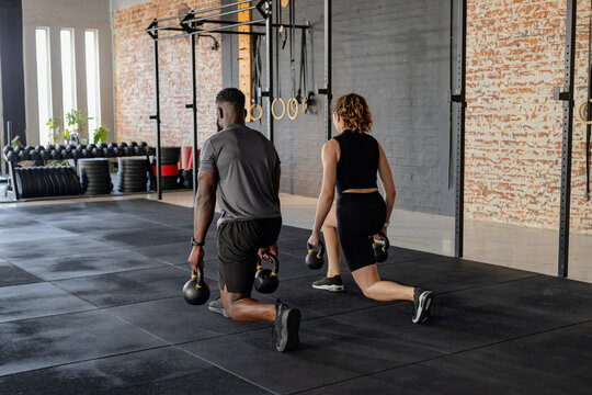 Diverse workout partners in athletic wear doing weighted lunges using kettlebells in CrossFit gym - Powered by Adobe