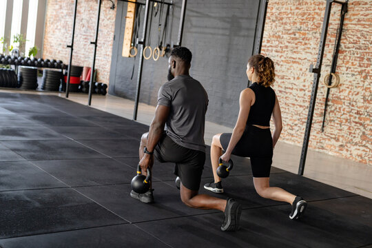 Diverse workout partners lunging in sportswear holding kettlebells in fitness studio with rings - Powered by Adobe