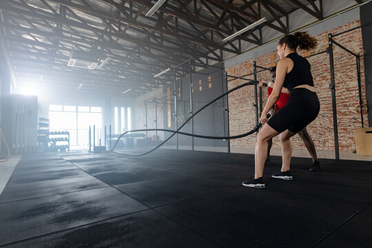 Workout partners woman and African American man using battle ropes at rig in gym, copy space