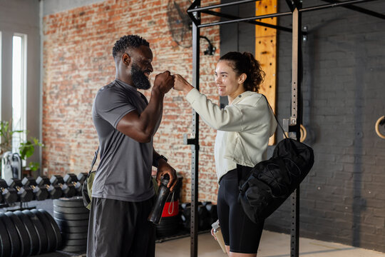 African American trainer and female client fist bumping in gym with dumbbells and weightlifting rig