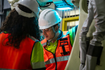 Two people wearing orange vests and hard hats. Team of engineers collaborating on programming and maintenance of a robotic arm in a smart factory.